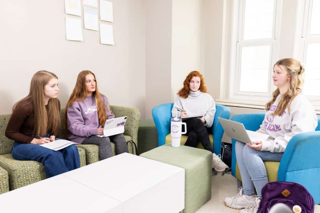 Four University of North Alabama students gather around for a meeting, laptops and notebooks in hand. They sit on a couch and two chairs.
