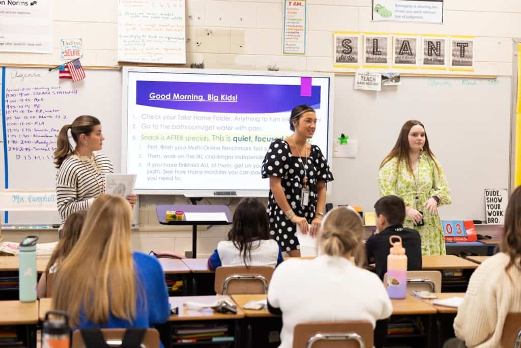 A group of three UNA student teachers stands at the front of a classroom. They're speaking to a classroom full of young students.