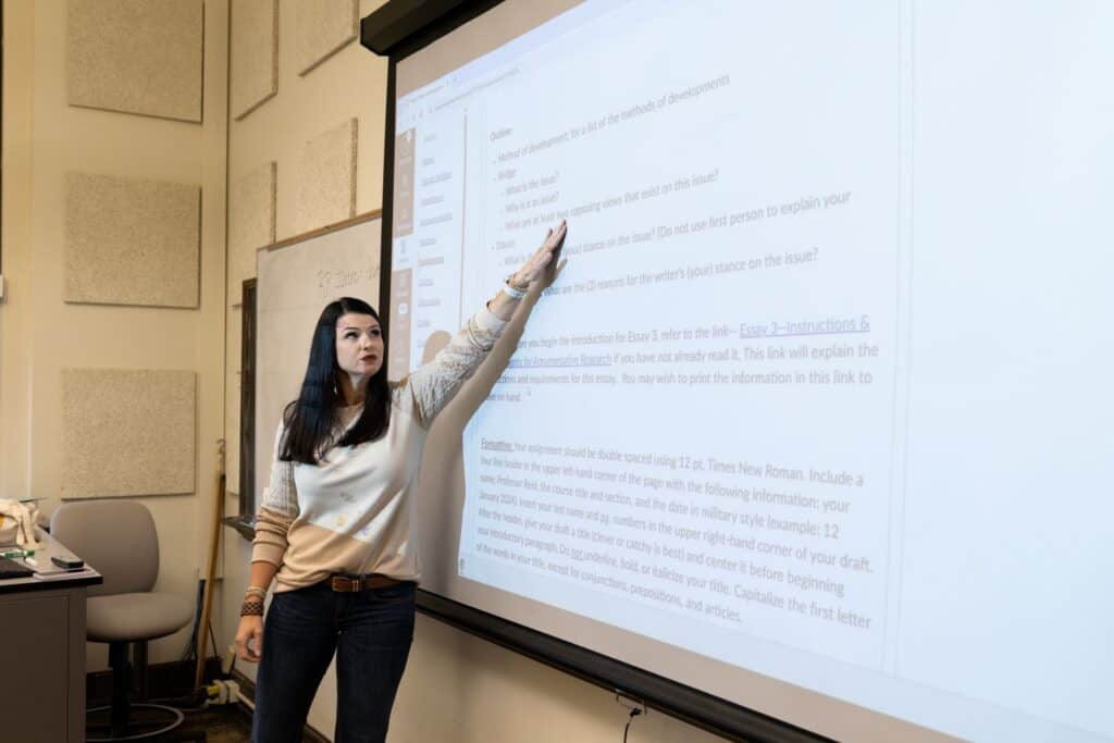 A University of North Alabama English professor stands at the front of a class. She points to a projector as she teaches.