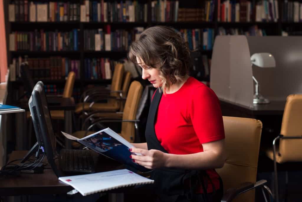 A woman reads a manuscript while sitting in a library.